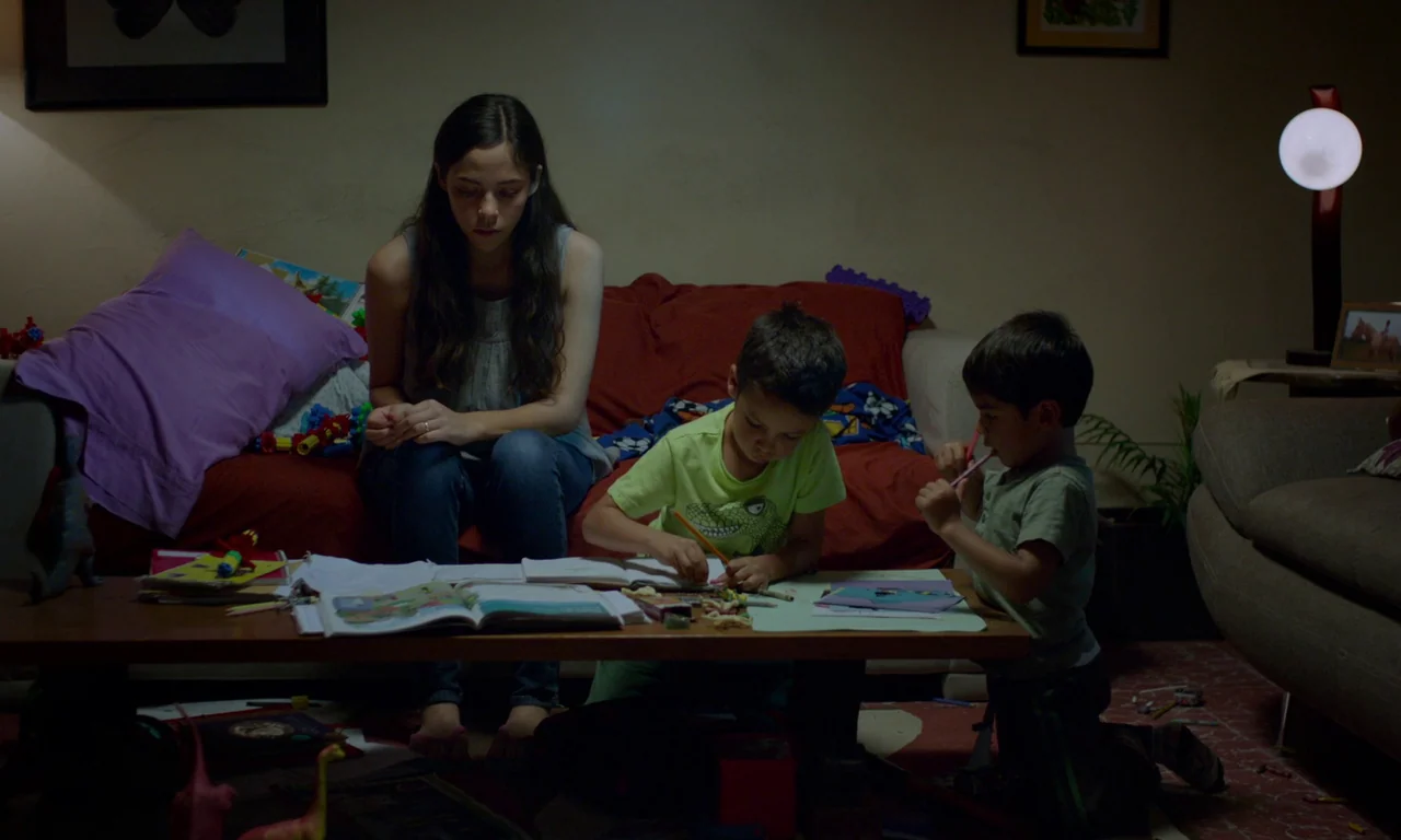 The Untamed (2016) movie still: medium — A dimly lit room contains a red sofa and a wooden coffee table cluttered with papers. A woman watches two…
