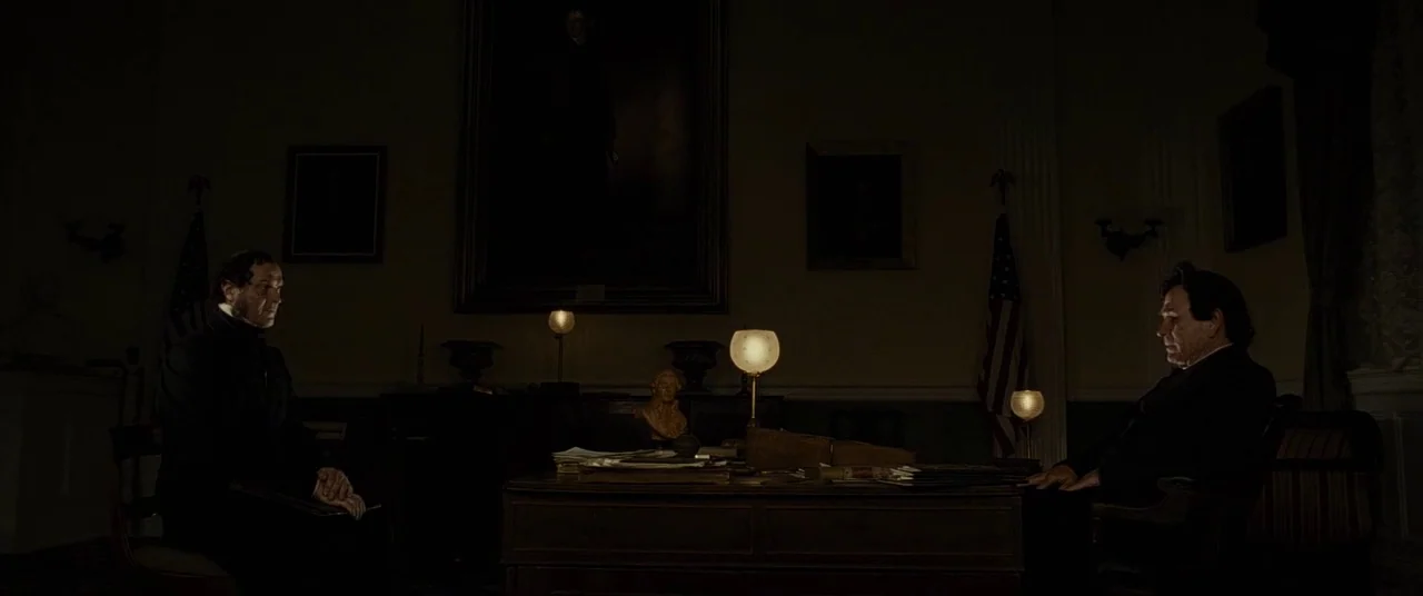 Lincoln (2012) movie still: wide — A dimly lit 19th-century presidential office features dark wood furniture and two US flags. Two men sit in chairs…