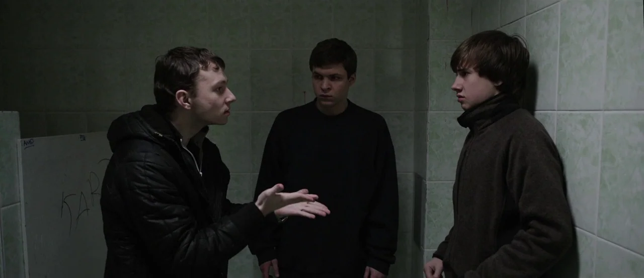 The Tribe (2014) movie still: medium — Three young men stand in a corner of a tiled public restroom facility. A man in a black jacket gestures as he…