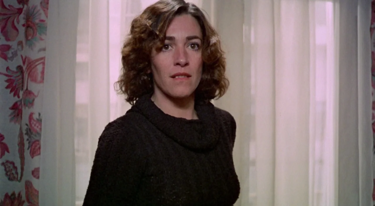 What Have I Done To Deserve This? (1984) movie still: medium — A woman stands indoors in front of a window covered by sheer, light-colored curtains.…