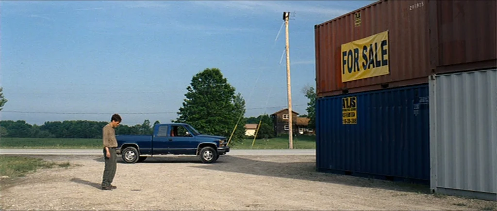 Take Shelter (2011) movie still: wide — A gravel lot sits under a clear blue sky next to stacked metal shipping containers. A young man stands alone…