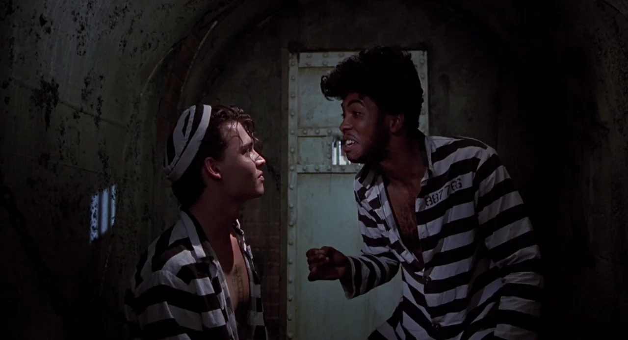 Cry Baby (1990) movie still: medium — A dimly lit, rounded stone prison cell features a heavy metal door. Two men in striped prisoner uniforms stand…