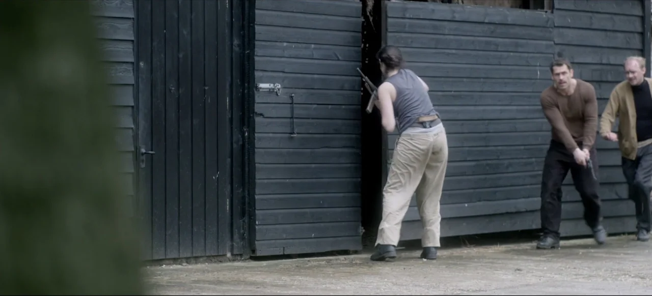 We Go In at Dawn (2020) movie still: fullbody — A dark wooden shed with horizontal planks stands on a concrete outdoor area. A woman in a tank top…