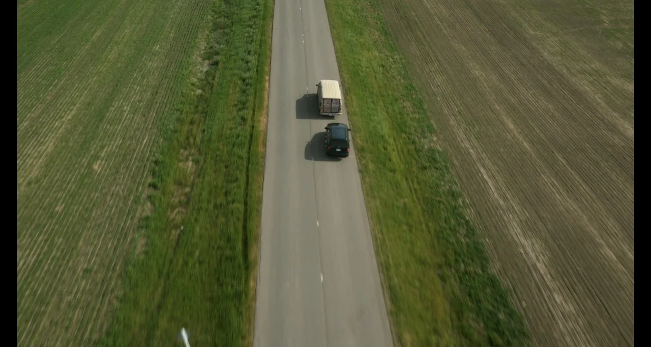 A.R.C.H.I.E. (2016) movie still: establishing — A straight asphalt road cuts through expansive grassy fields and tilled agricultural land. A dark…