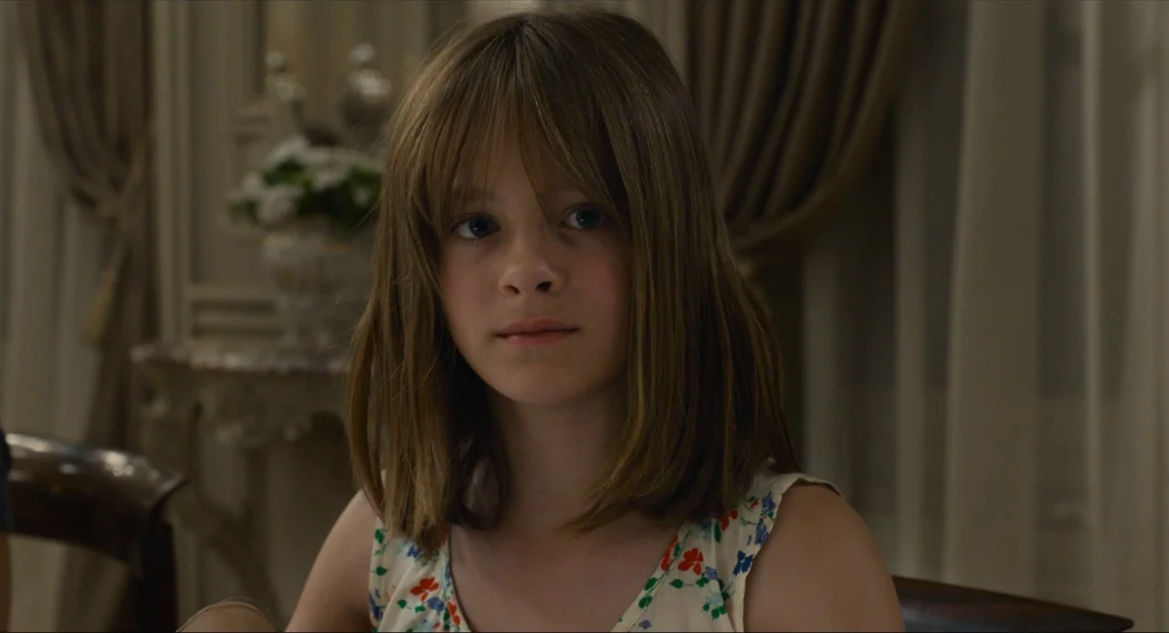 Happy End (2017) movie still: closeup — A young girl sits in a brightly lit dining area with curtains visible. The girl looks forward with a neutral…