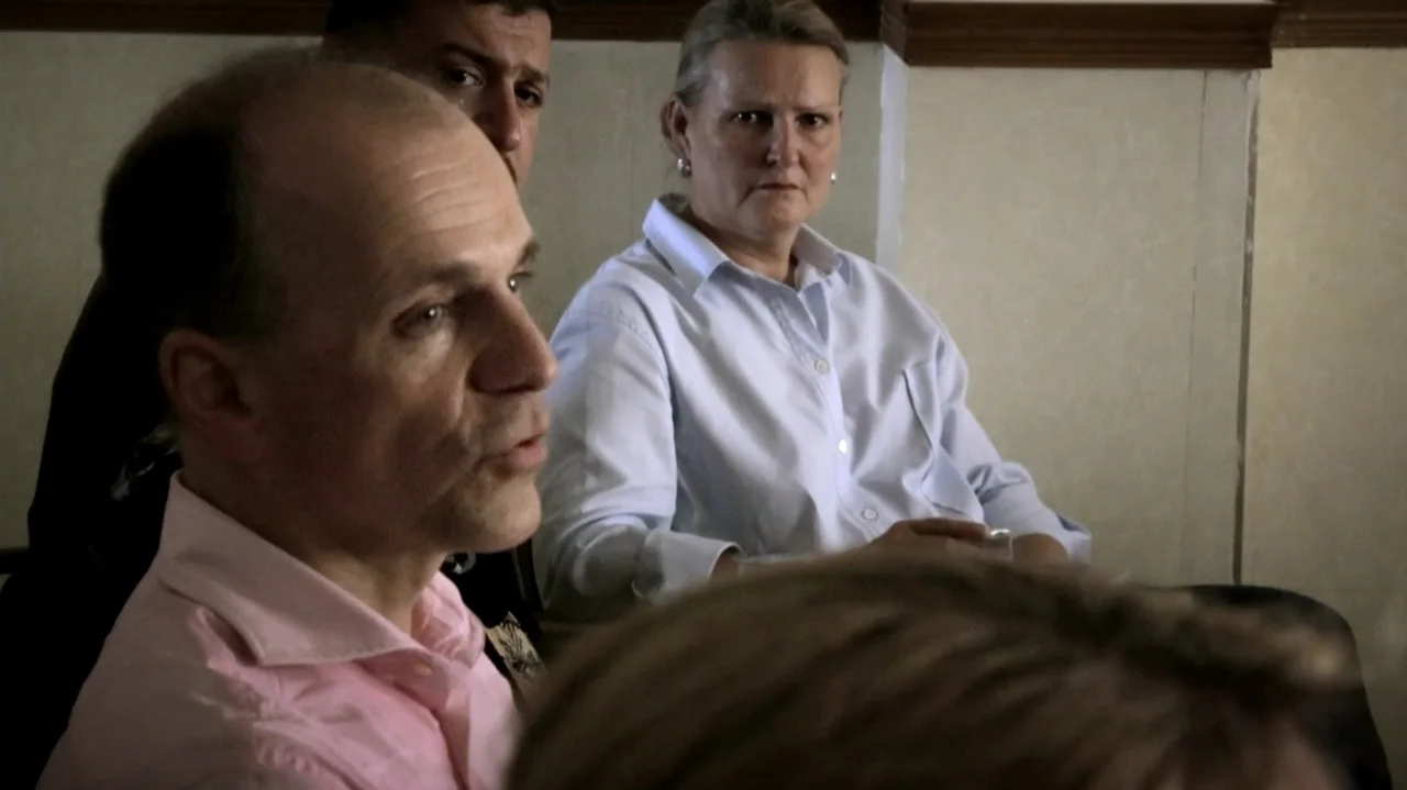 City of Two Springs (2019) movie still: medium — A plain interior room with cream-colored walls and a wooden trim at the ceiling. Three adults sit…