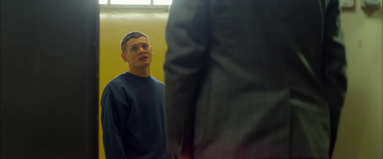 Starred Up (2013) movie still: medium — A stark indoor corridor features mustard-yellow walls and a high rectangular window. A young man in a blue…