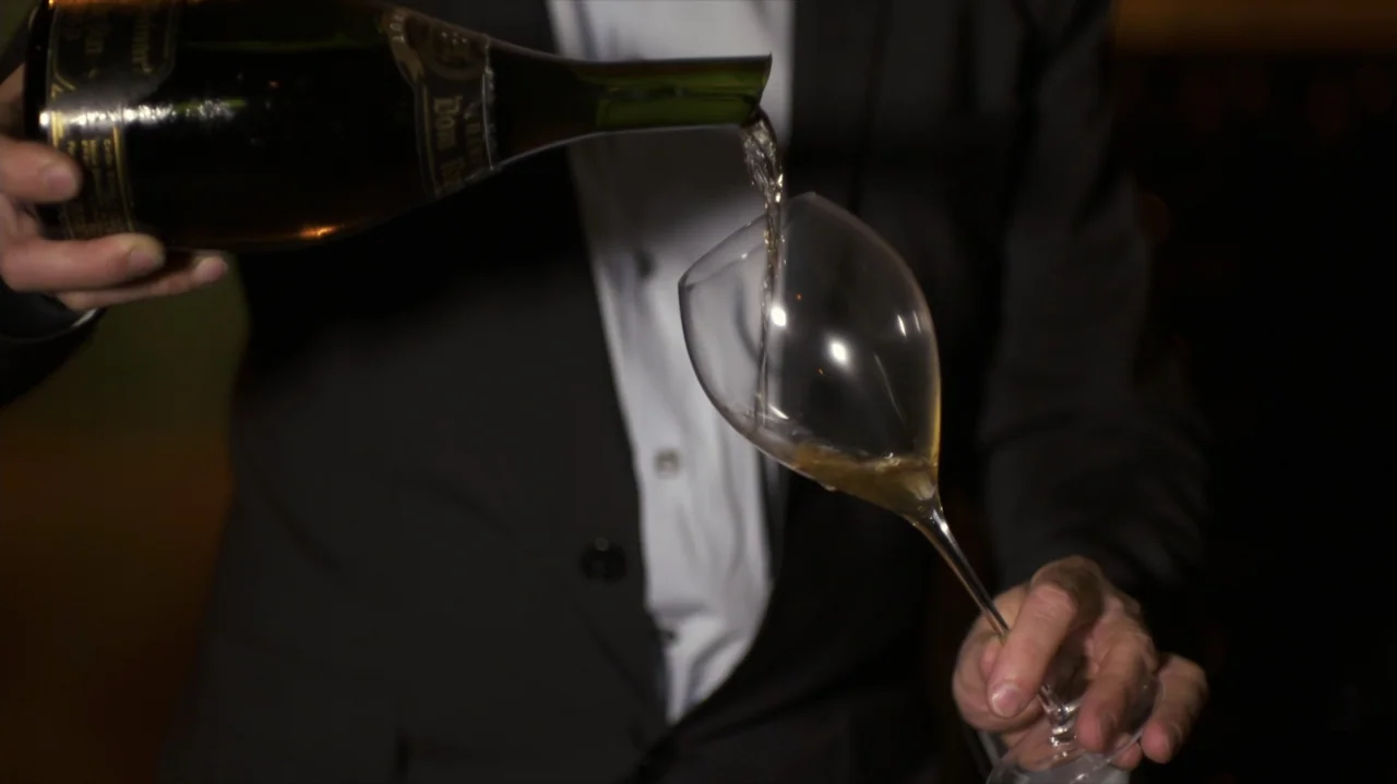 Somm: Into the Bottle (2015) movie still: medium — A dimly lit indoor space features a subject in formal wear. A person pours champagne from a glass…