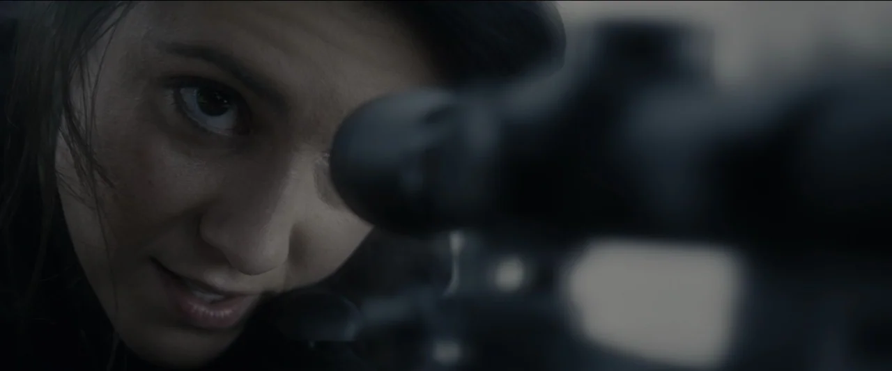 Kate (2021) movie still: closeup — A woman aims a rifle on an outdoor rooftop against a blurred urban backdrop. She peers intensely through the…