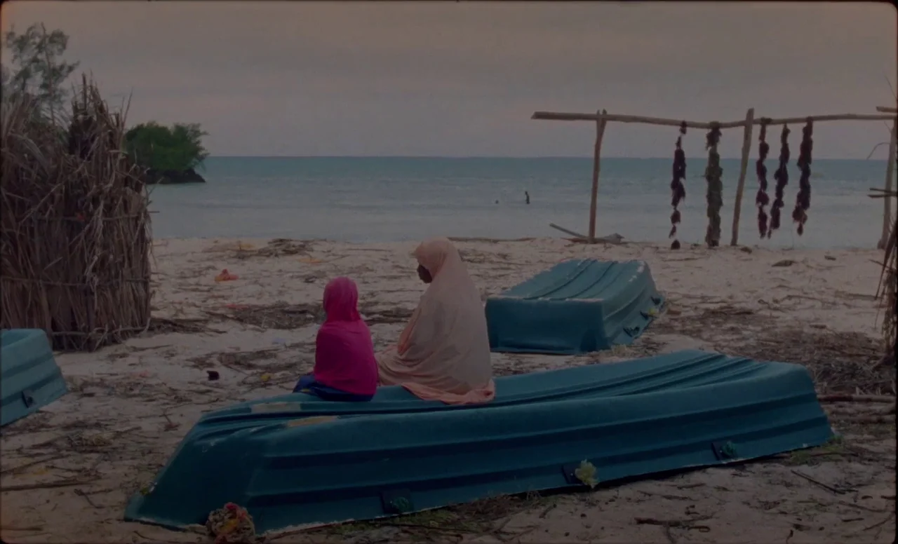 Samsara (2023) movie still: wide — A sandy beach at dusk features two people sitting on overturned blue plastic boats. An improvised wooden rack…