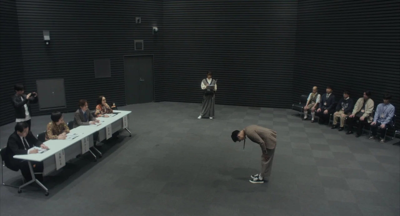 Stigmatized Properties: Possession (2025) movie still: wide — A spacious room with dark slatted walls and a grey carpeted floor. A man bows in the…