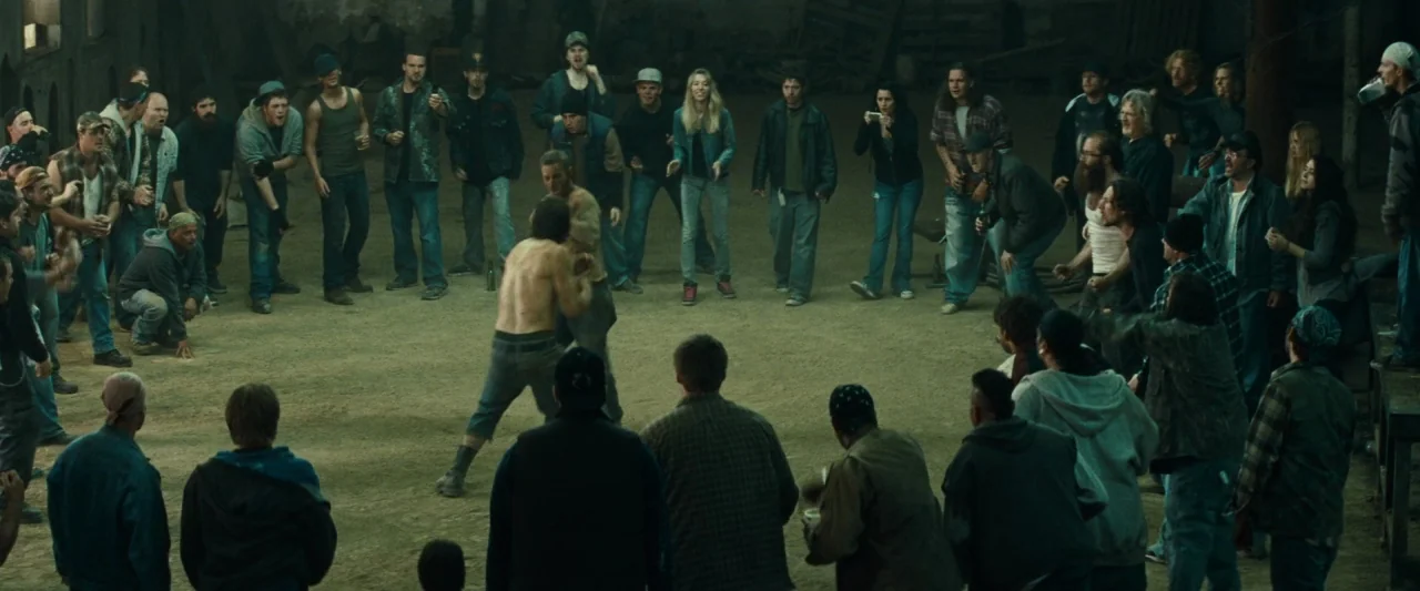 Out of the Furnace (2013) movie still: wide — A dusty, cavernous industrial space is filled with a large crowd of people. Two men are engaged in a…