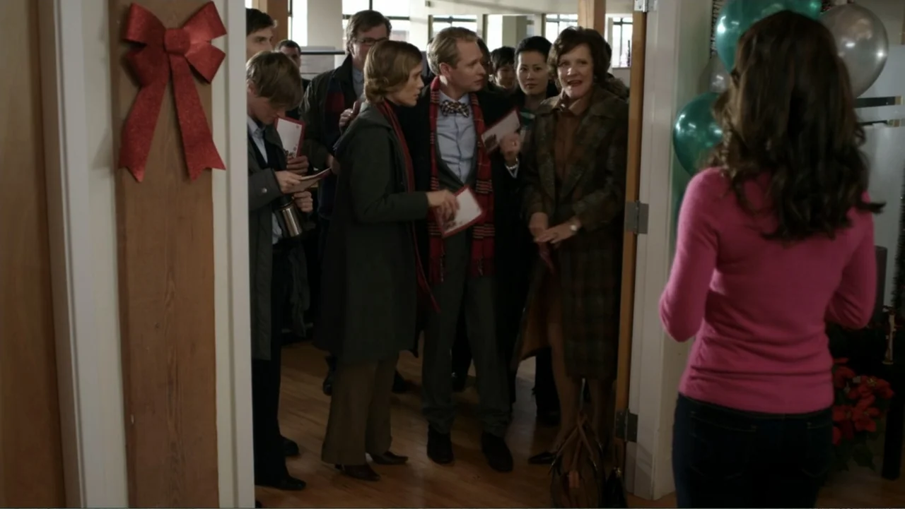 It's Christmas, Carol! (2012) movie still: medium — An office doorway contains a group of people holding cards at a holiday event. A woman in a pink…