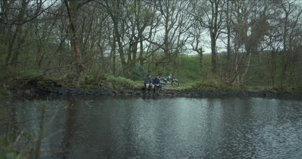 God’s Own Country (2017) movie still: wide — A quiet riverbank edged by a stone wall sits before a dense, leafless forest. Two men sit together on…