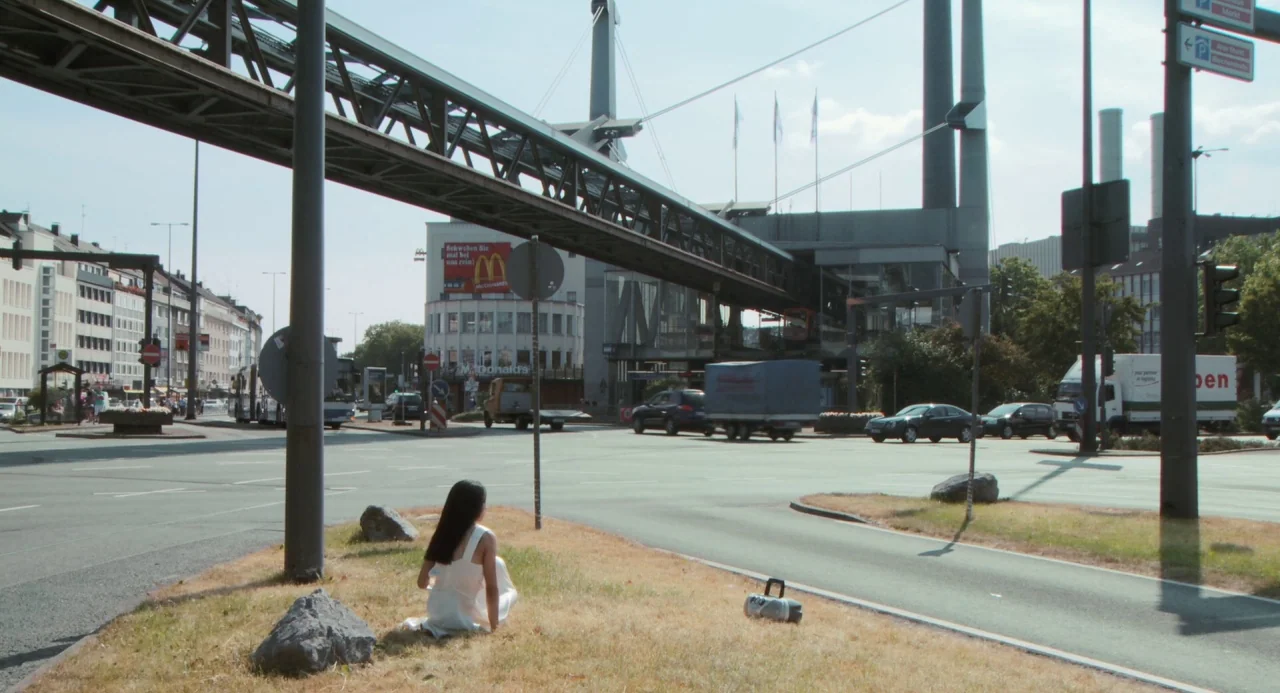 Pina (2011) movie still: wide — A wide urban intersection features a prominent elevated monorail track overhead during the daytime. A lone woman…