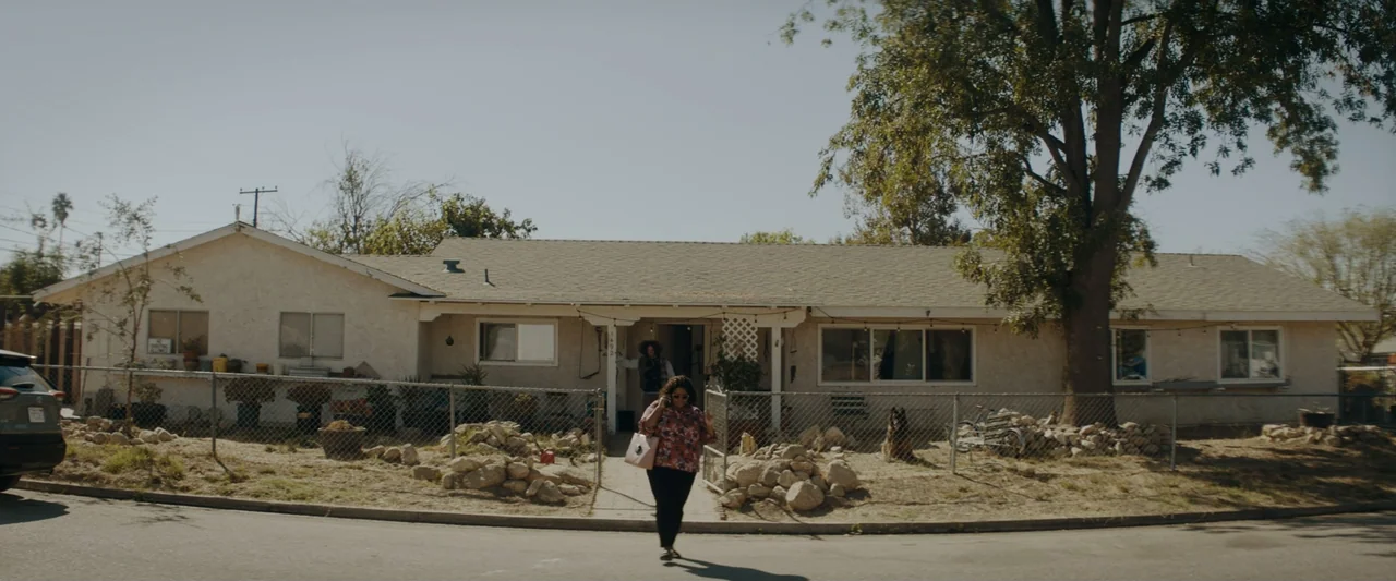 Encounter (2018) movie still: wide — A single-story suburban house sits behind a chain-link fence with a large tree. A woman walks along the asphalt…