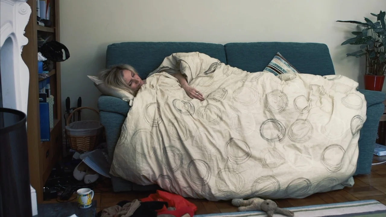 You Might Get Lost (2021) movie still: medium — A living room interior featuring a blue sofa and a houseplant in a red pot. A woman with blonde hair…