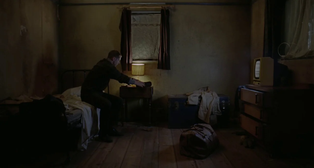 Possum (2018) movie still: medium — A dimly lit, cramped room contains a bed, a wooden trunk, and a small television. A man sits on the side of the…