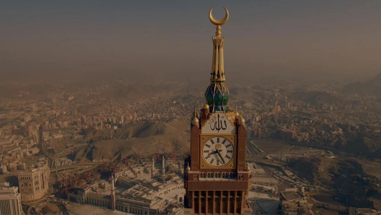One Day in the Haram (2017) movie still: establishing — A high-angle view shows the top of a monumental clock tower in Mecca. The surrounding desert…
