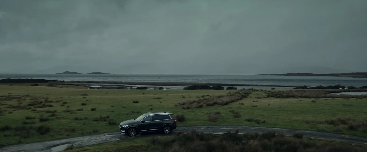 My Son (2021) movie still: wide — A dark grey SUV travels along a narrow asphalt path through a coastal field. The background consists of a grey…