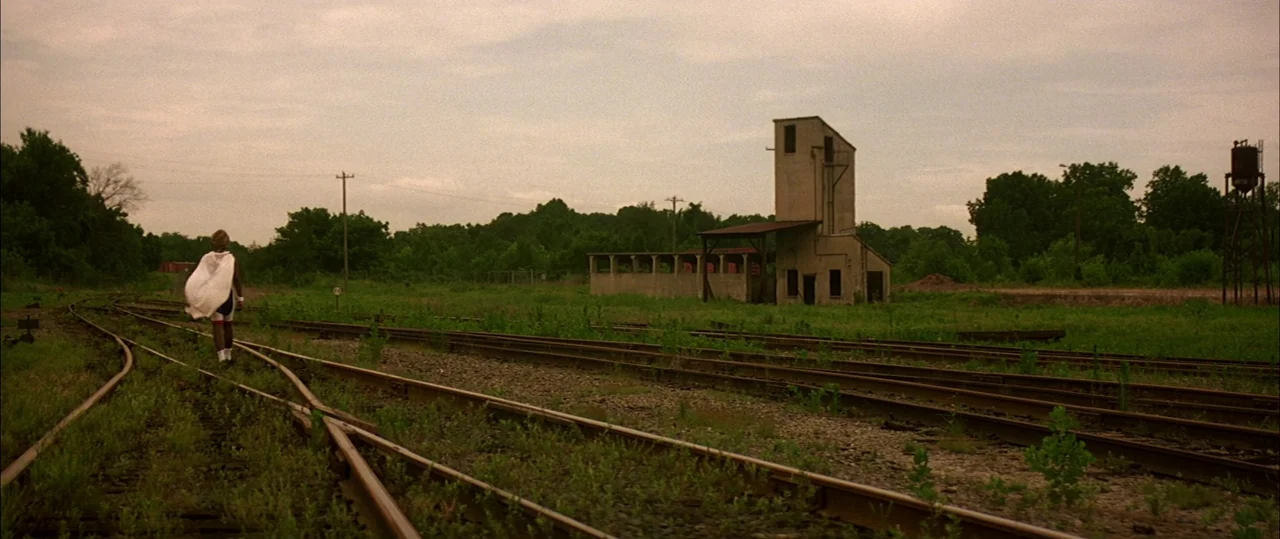 George Washington (2000) movie still: wide — A rural landscape features overgrown railway tracks leading toward a concrete industrial structure. A…