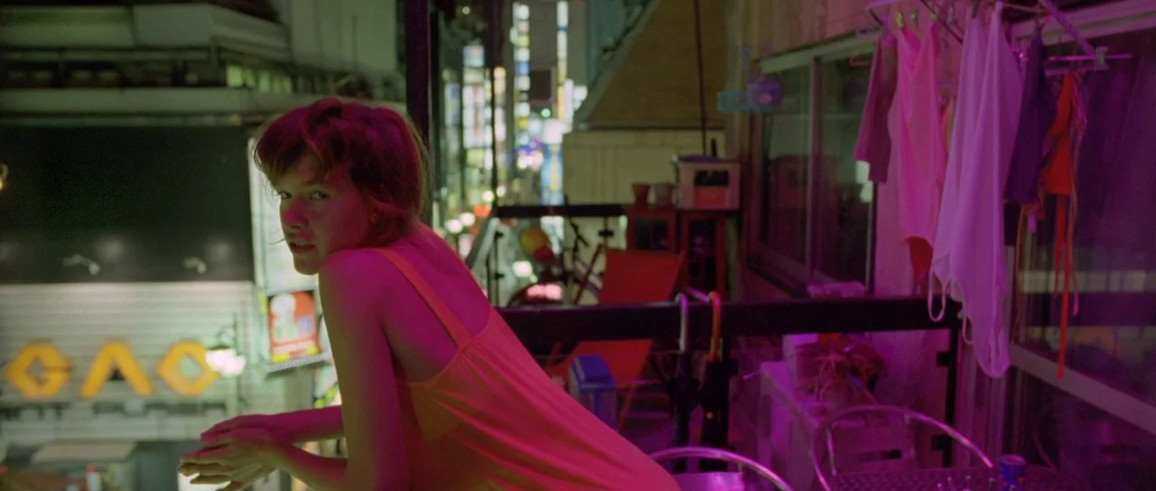 Enter The Void (2009) movie still: medium — An urban apartment balcony at night illuminated by glowing signs from the street below. A woman leans on…