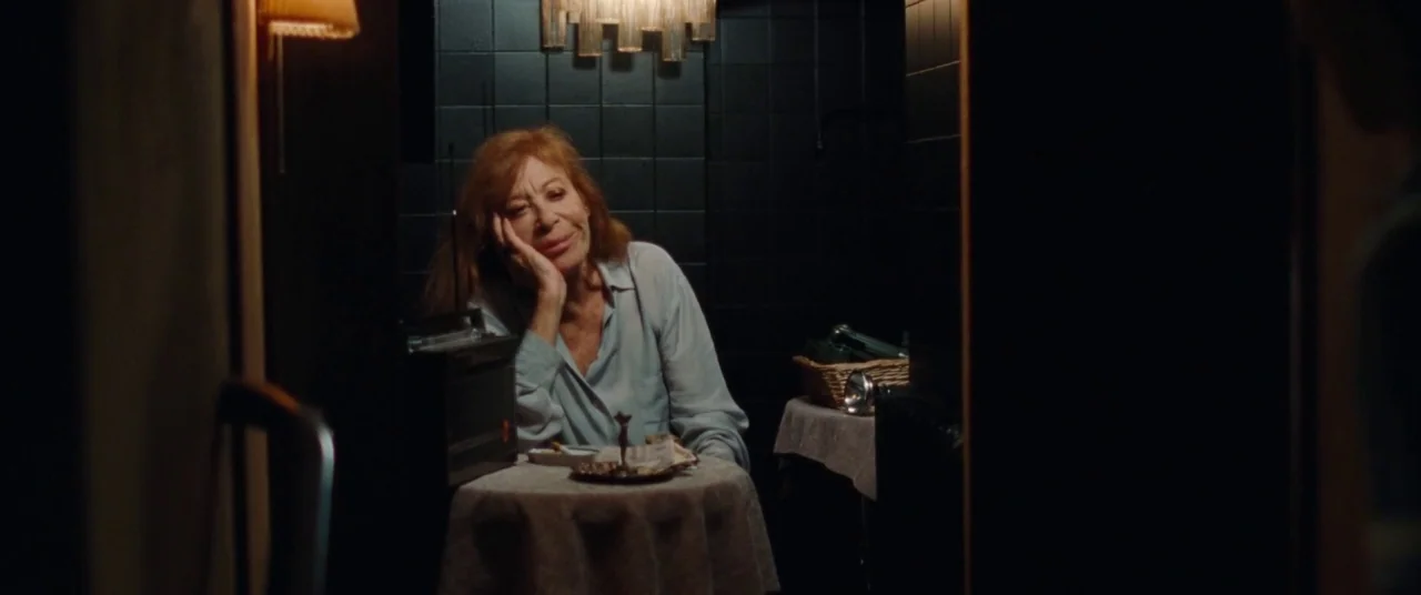 Knife+Heart (2018) movie still: medium — A dimly lit room features black tiled walls and a small table with a radio. An older woman sits at the…