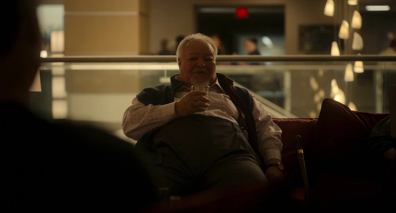 Civil War (2024) movie still: medium — A dark hotel lobby features a seating area with red upholstered furniture. A man in a sweater vest sits on a…