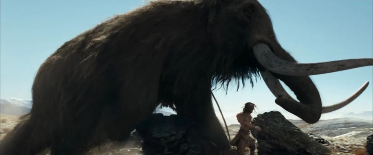 10,000 BC (2008) movie still: wide — An arid, rocky landscape features sparse vegetation under a clear, bright sky. A man runs near a massive woolly…
