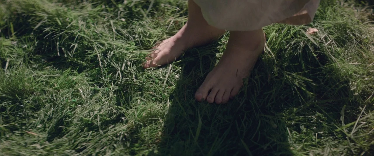The Dancer movie still: closeup — A patch of wild green grass is textured with diverse blades and small weeds. A person stands barefoot on the…