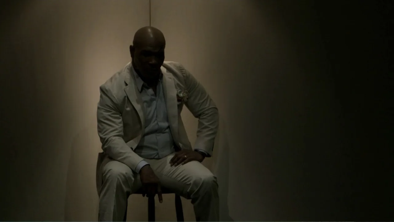Mike Tyson: Undisputed Truth (2013) movie still: medium — A man sits on a stool against a plain, dimly lit corner wall. He is wearing a beige suit…