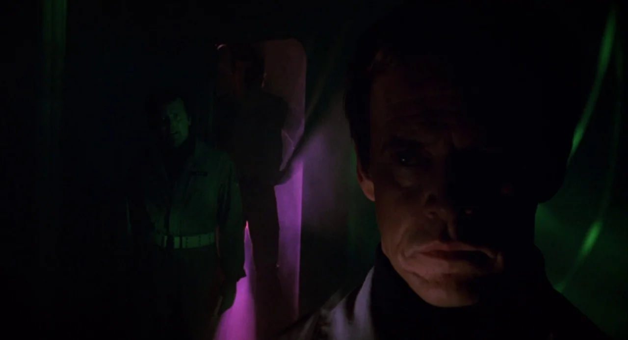 Night of the Comet movie still: closeup — A dimly lit interior space contains metallic wall panels and a bright neon-lit doorway. One man occupies…