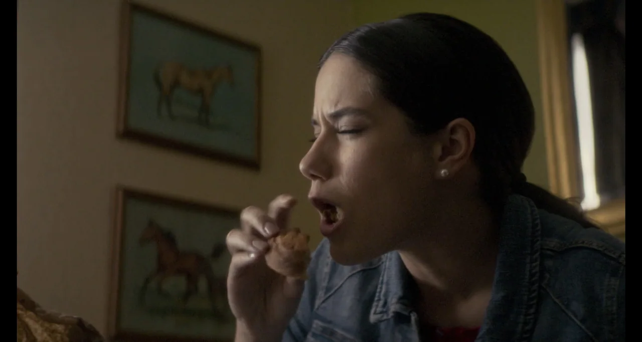 The Poison Rose (2019) movie still: closeup — An interior room has beige walls decorated with framed horse prints. A woman in a denim jacket eats a…