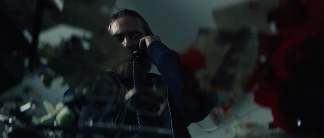 You Were Never Really Here (2017) movie still: medium — A dimly lit room with out-of-focus objects in the immediate foreground. A bearded man speaks…