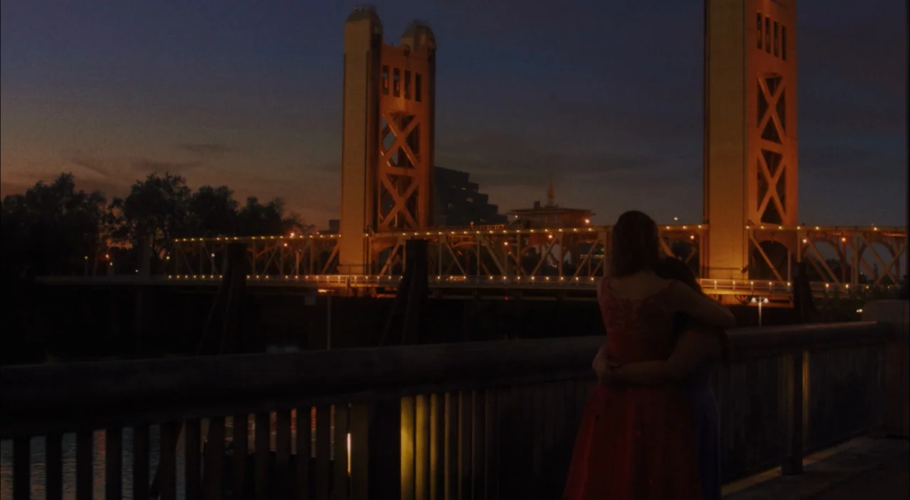 Lady Bird (2017) movie still: wide — A nighttime view of a large vertical lift bridge illuminated by amber street lighting. Two women embrace on a…