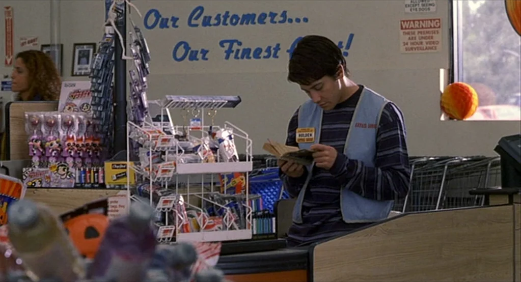 The Good Girl (2002) movie still: medium — A brightly lit retail checkout area features a worker behind a wooden counter. A young man stands behind…