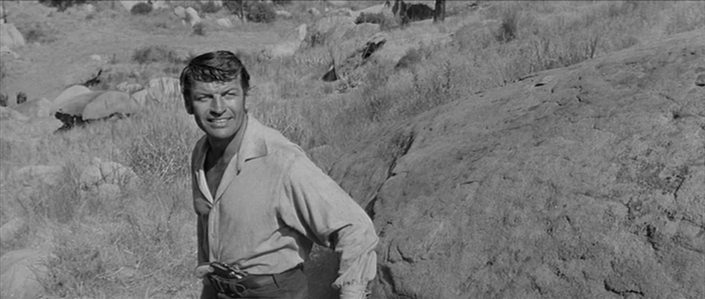 Love Me Tender movie still: medium — A rugged landscape filled with dry grasses and scattered large granite boulders. A man in a simple button-up…
