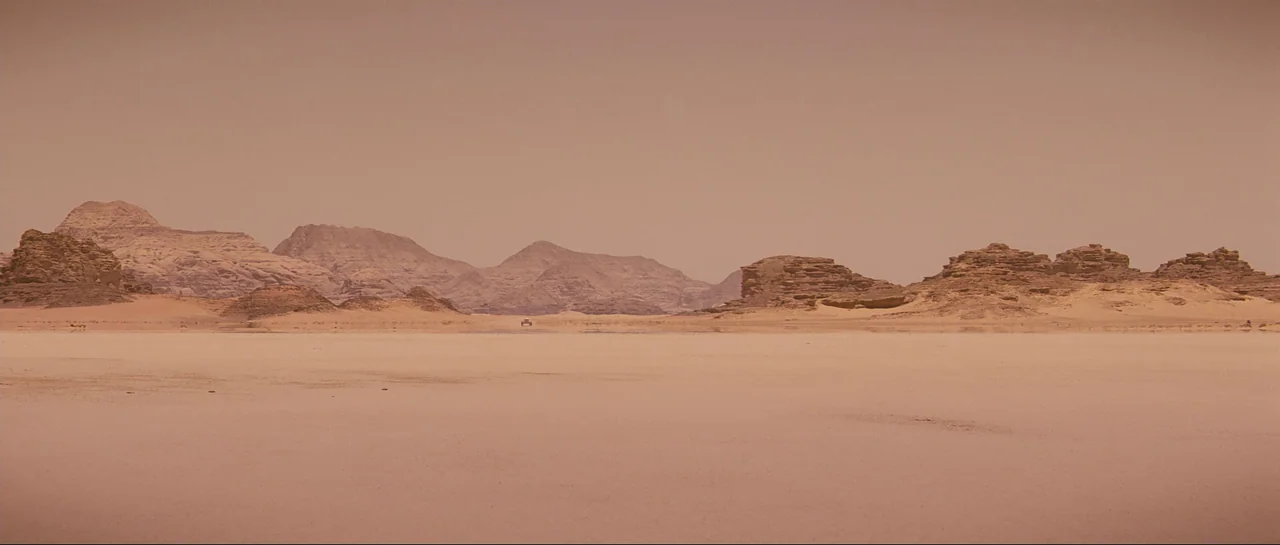 The Last Days On Mars (2013) movie still: establishing — A vast, flat desert basin with rugged rocky formations lining the distant horizon. A tiny…
