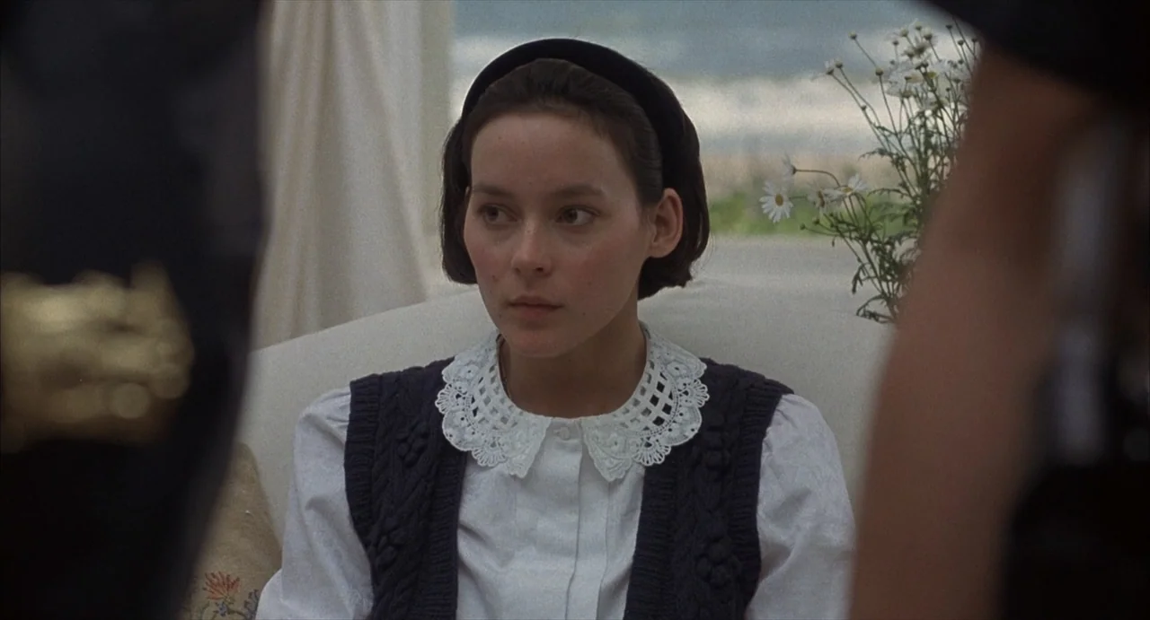 Masquerade (2023) movie still: medium — A light-filled sunroom features white upholstery and a vase of daisies by a window. A woman wearing a knit…