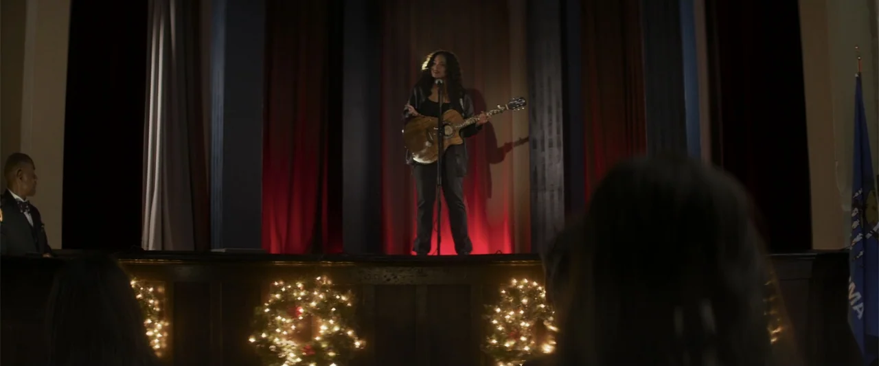 Holiday Harmony (2022) movie still: fullbody — A wooden stage features a musician performing before an audience in a darkened room. The stage has…