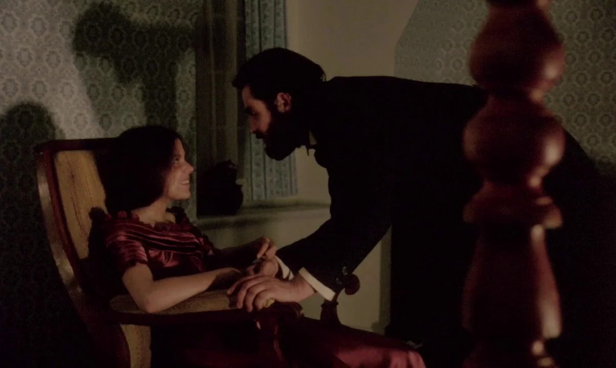 Kamouraska (1973) movie still: medium — A dimly lit room with patterned wallpaper features a woman seated in a chair. A man wearing a black suit…