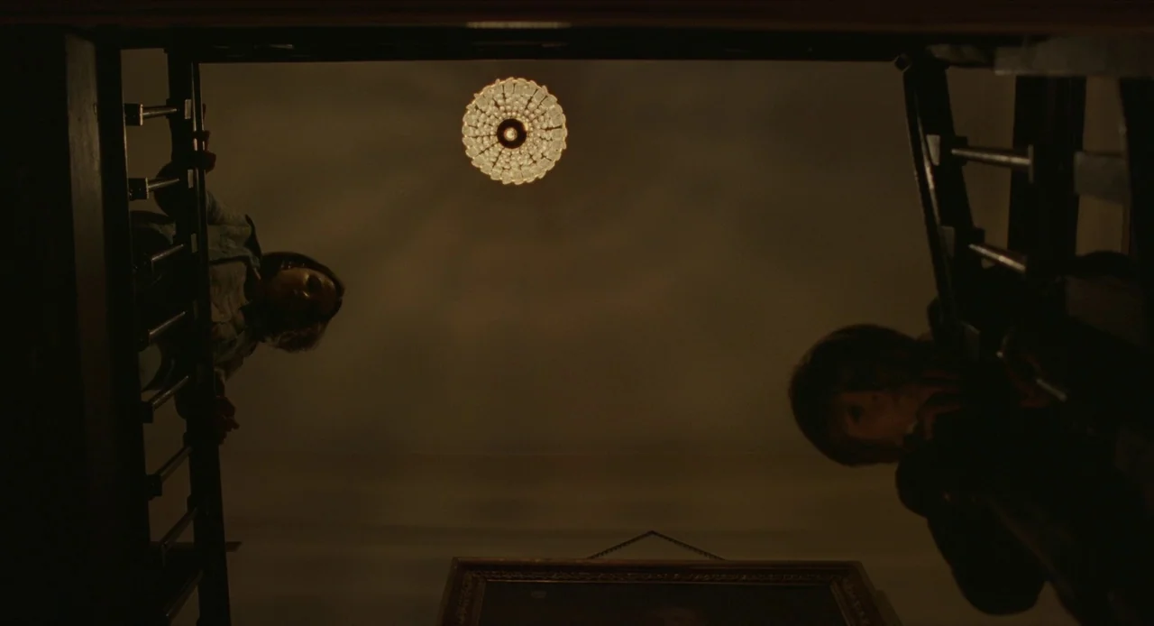 The Nest (2020) movie still: wide — A view looking up from the bottom of a dark stairwell toward the landing. Two figures lean over a wooden…