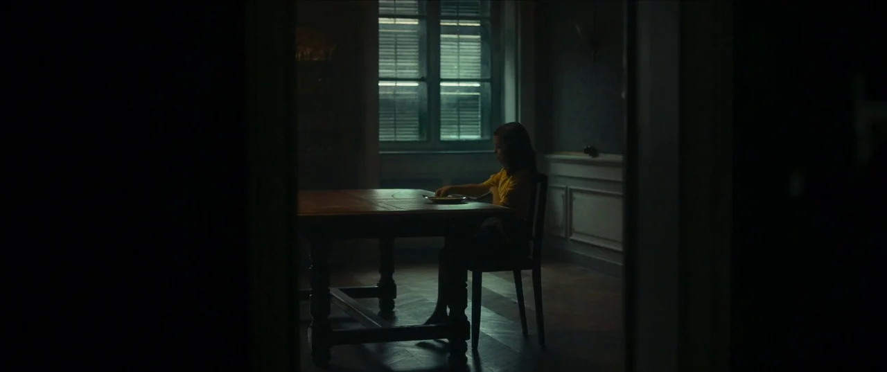 Earwig (2022) movie still: wide — A dark dining room features a heavy wooden table and a window with blinds. A young girl sits alone at the table…