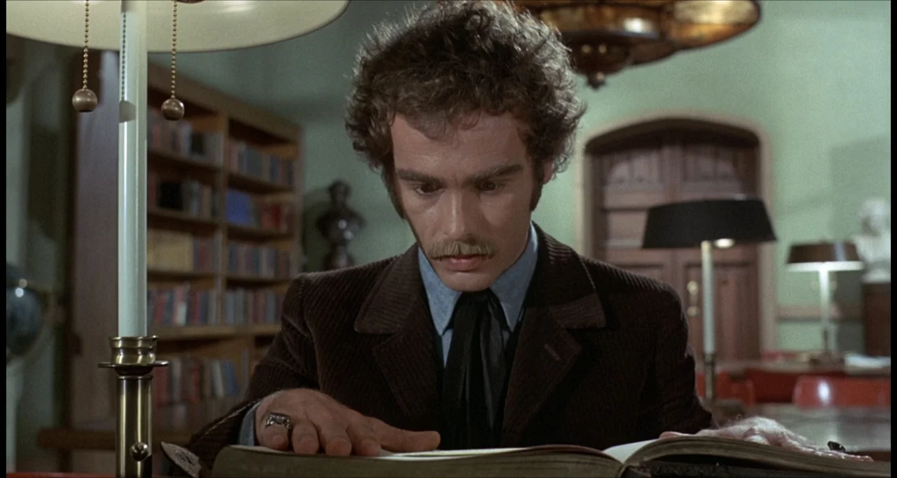 The Dunwich Horror (1970) movie still: medium — A quiet room filled with shelves of books and classic lamp fixtures. A young man sits at a desk…