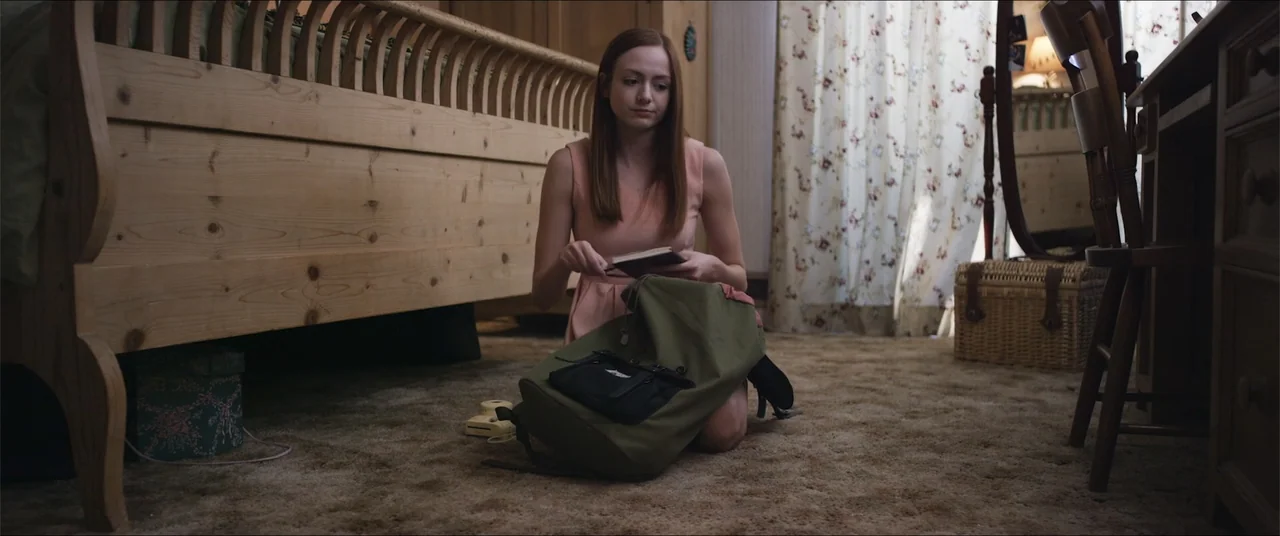 Captive (2020) movie still: medium — A bedroom contains a light wooden bed frame and a patterned floral curtain background. A young woman with…