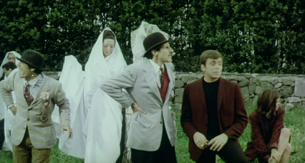 Dr. Goldfoot and the Girl Bombs (1966) movie still: fullbody — A group of people stand and sit outdoors near a low dry-stone wall. Thick dark bushes…
