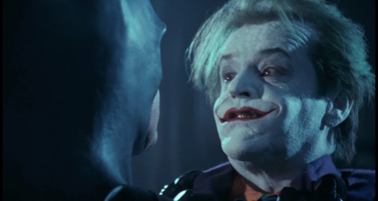 Batman (1989) movie still: medium — A dark indoor space contains two men positioned close to one another in shadow. A man with white face paint and…