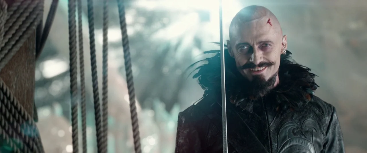 Pan (2015) movie still: medium — The deck of a sailing ship features thick hanging ropes and blurred background structures. A bald man wearing a…
