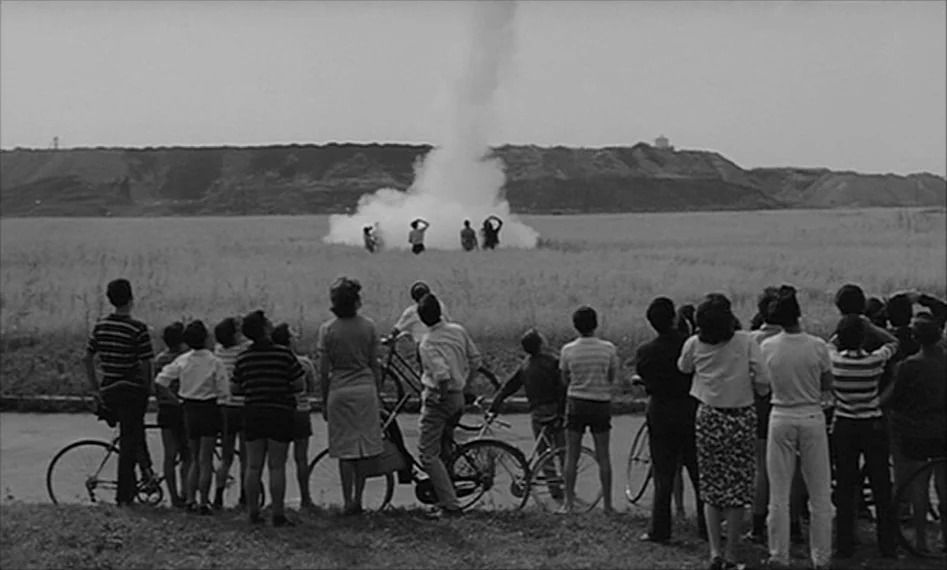 La Notte (1961) movie still: wide — A group of people stands along a road observing smoke in a flat field. Beyond the field, a low earthen ridge…