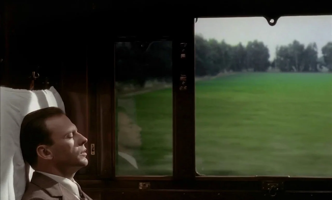 The Conformist (1970) movie still: medium — A wooden train compartment interior features a window with a view of green fields. A man in a suit rests…